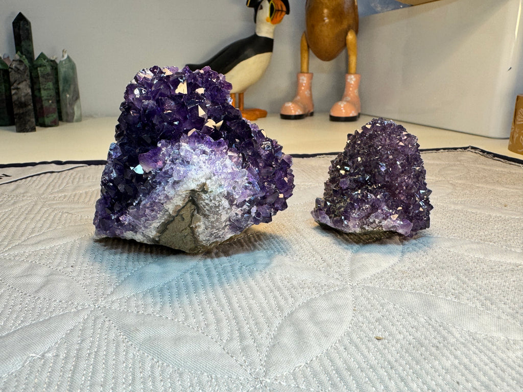 Two purple amethyst geodes on a textured surface with decorative items in the background.