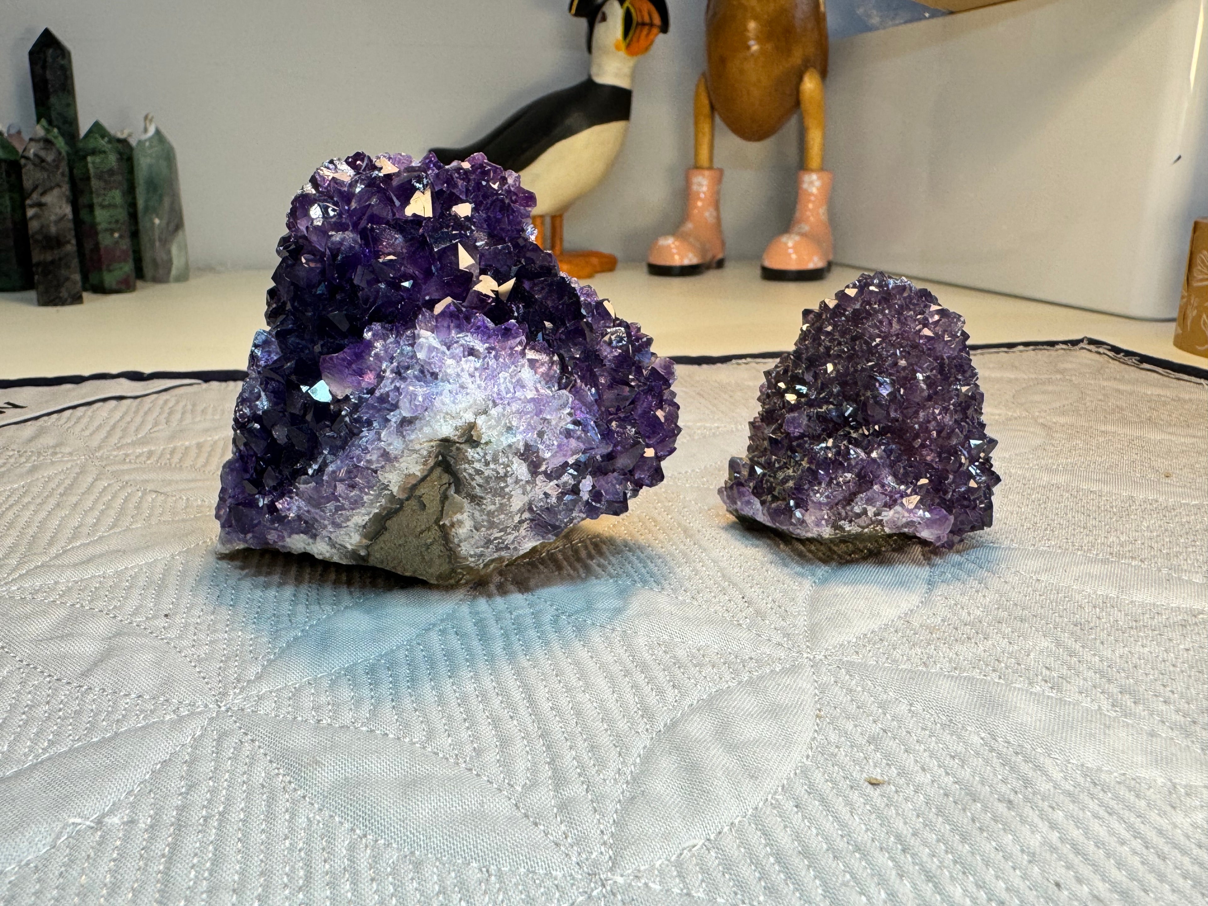 Two purple amethyst geodes on a textured surface with decorative items in the background.