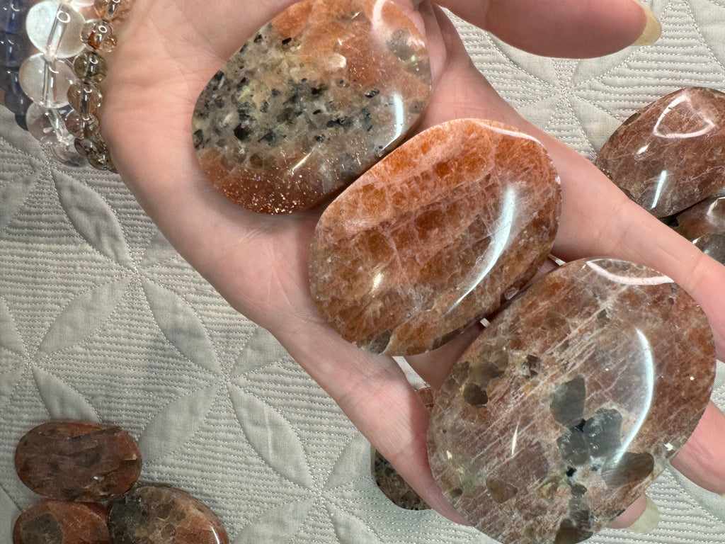 Hand holding a polished brown stone with more stones in the background on a textured surface