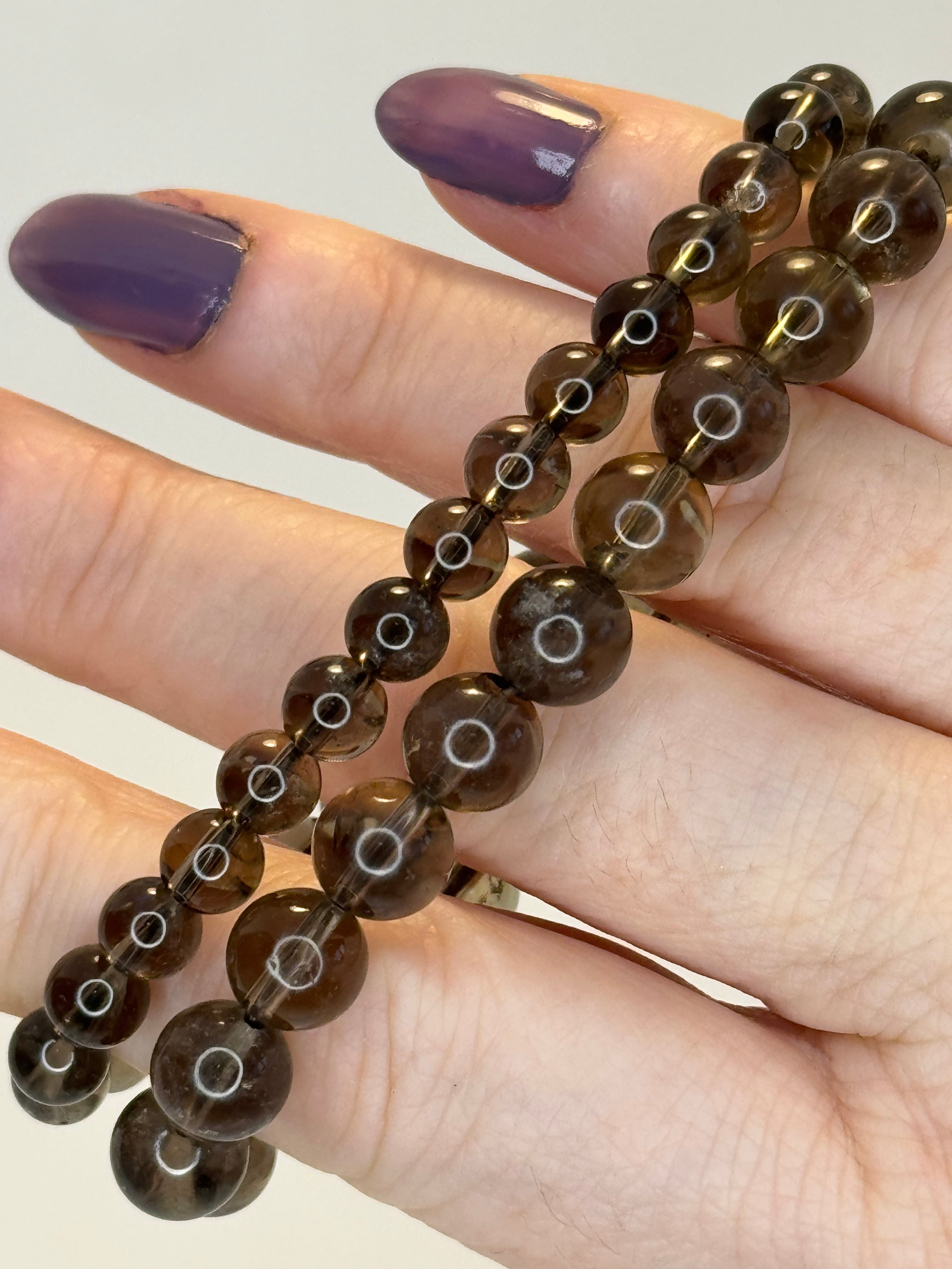 2 Brown beaded bracelets on a hand with purple nail polish against a neutral background