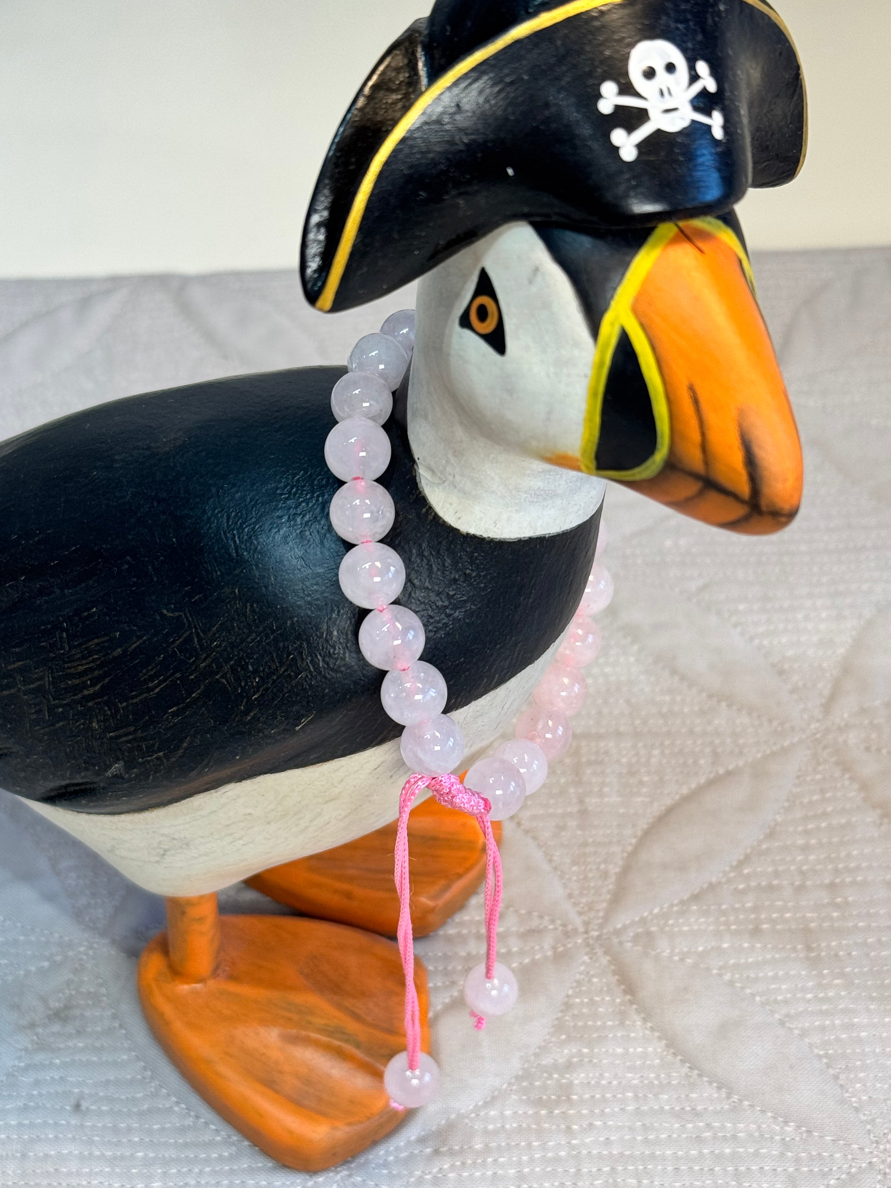 Puffin figurine wearing a pirate hat with beaded rose quartz bracelet on a textured surface