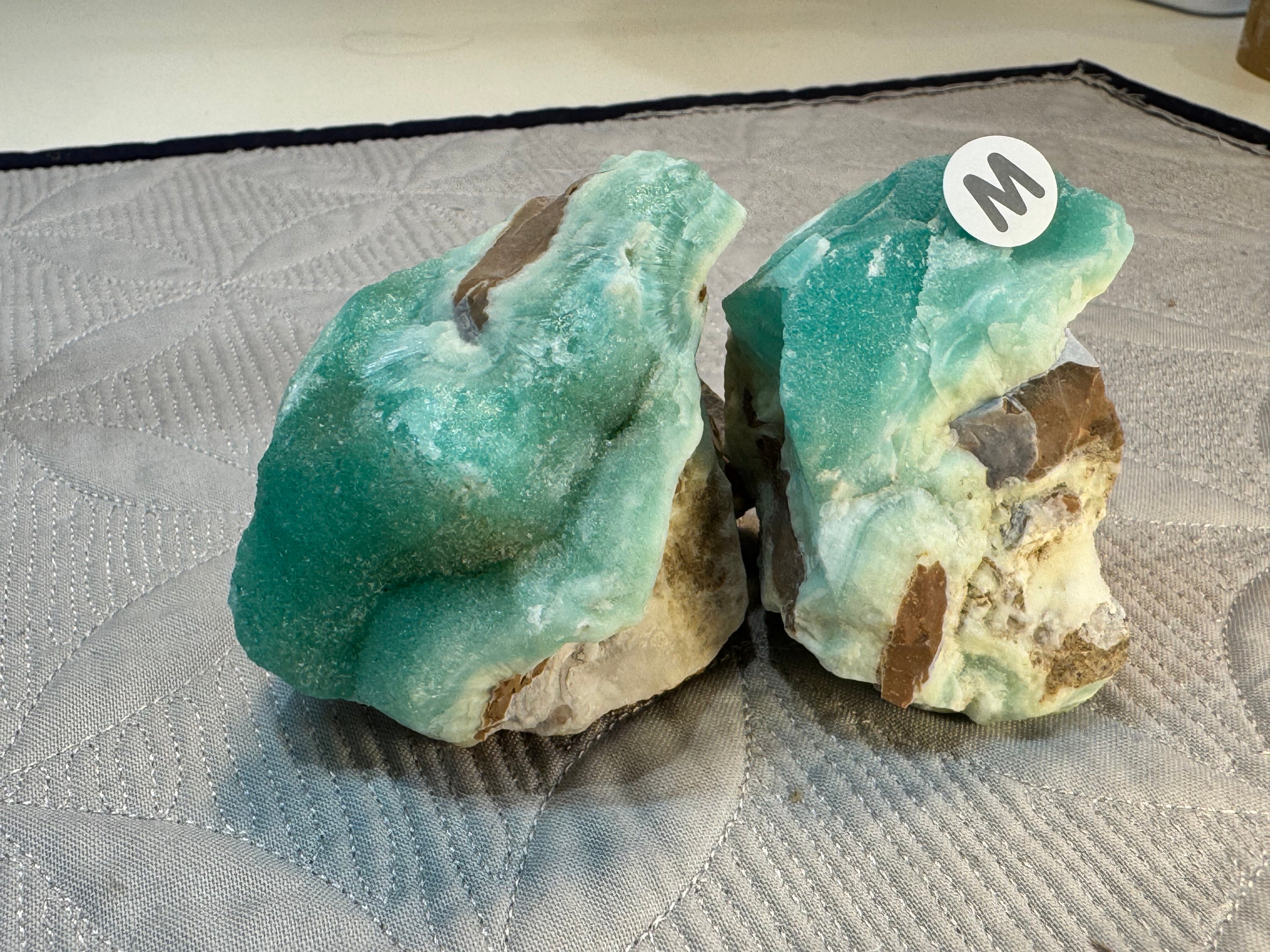 Two turquoise-colored rocks on a textured surface with a brand logo in the corner.