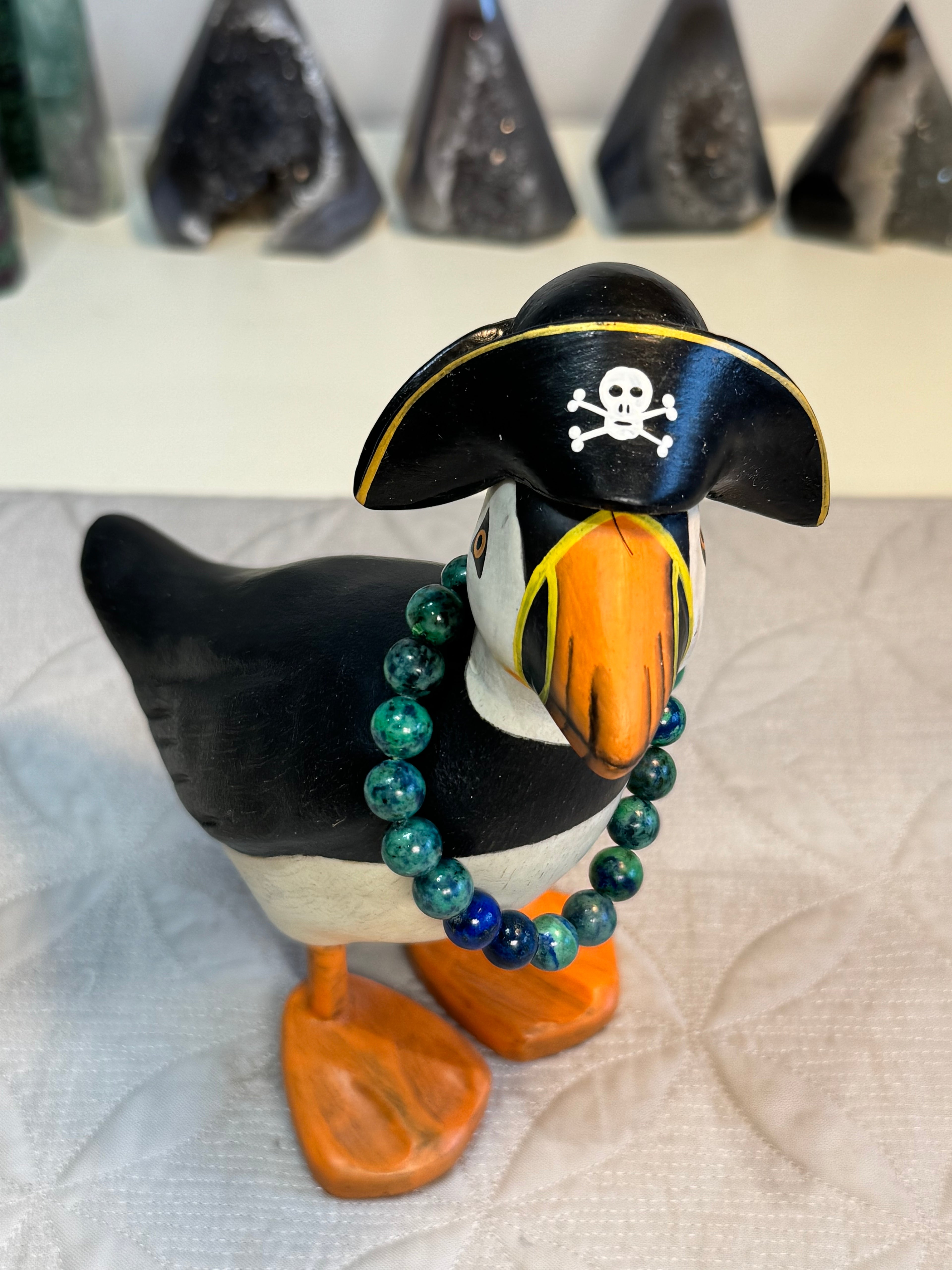 Puffin figurine wearing a pirate hat with a skull and crossbones on a textured white surface. with bracelet around its neck