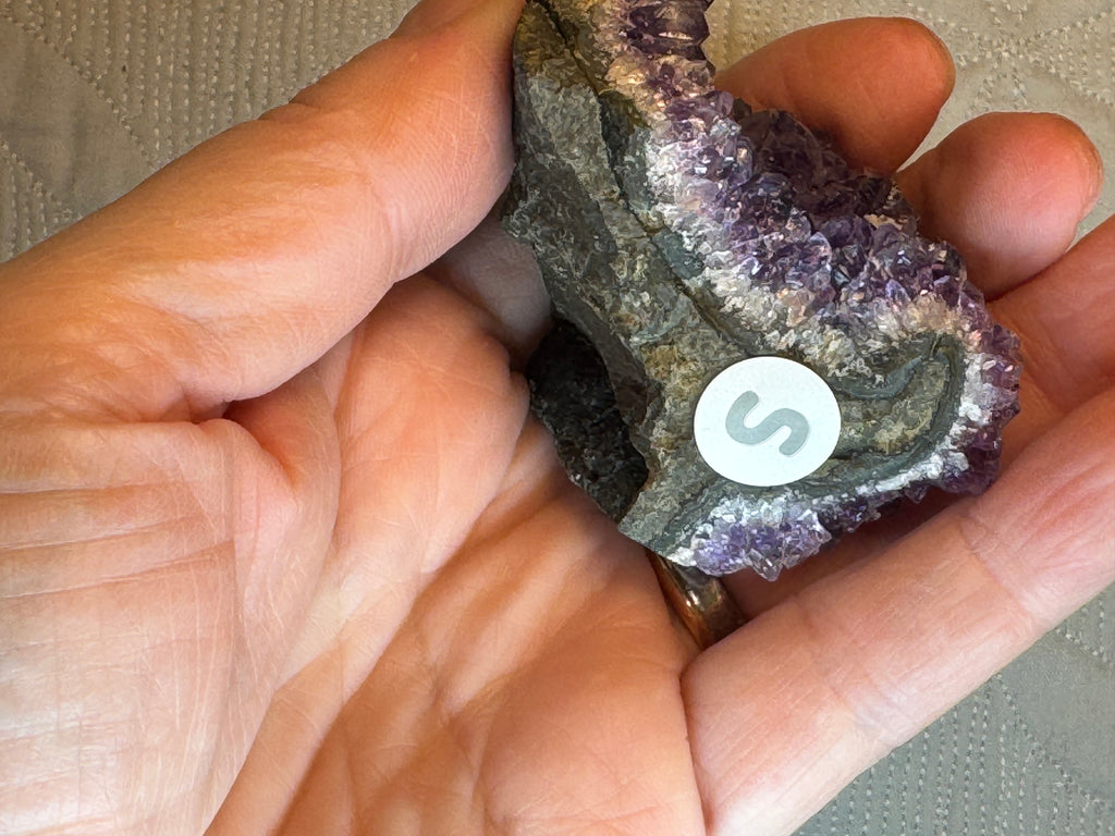 Hand holding a crystal with a 'S' symbol, against a neutral background