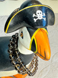 Puffin figurine wearing a pirate hat with a skull and crossbones on a textured white surface. with 2 bracelets around its neck