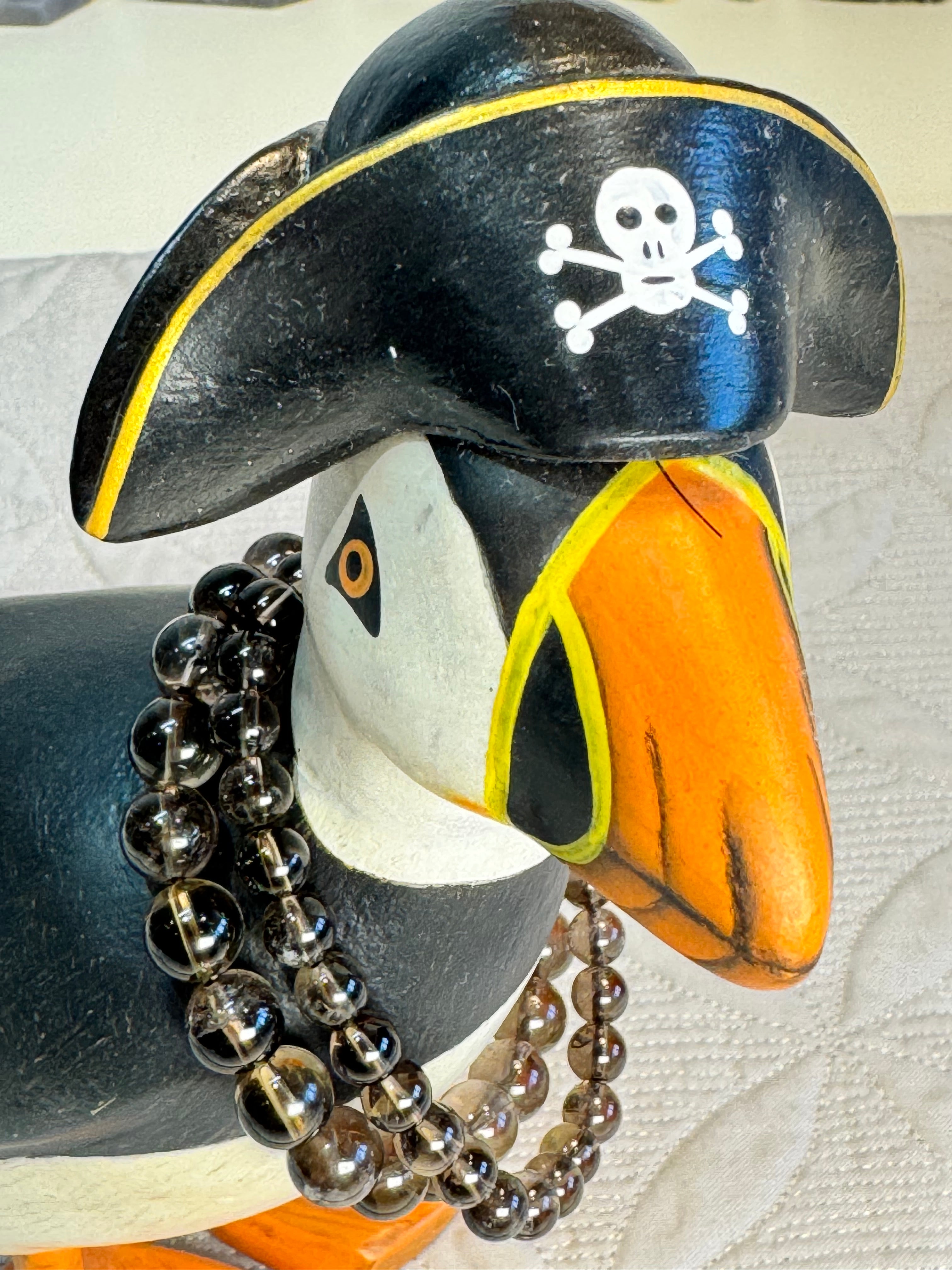Puffin figurine wearing a pirate hat with a skull and crossbones on a textured white surface. with 2 bracelets around its neck