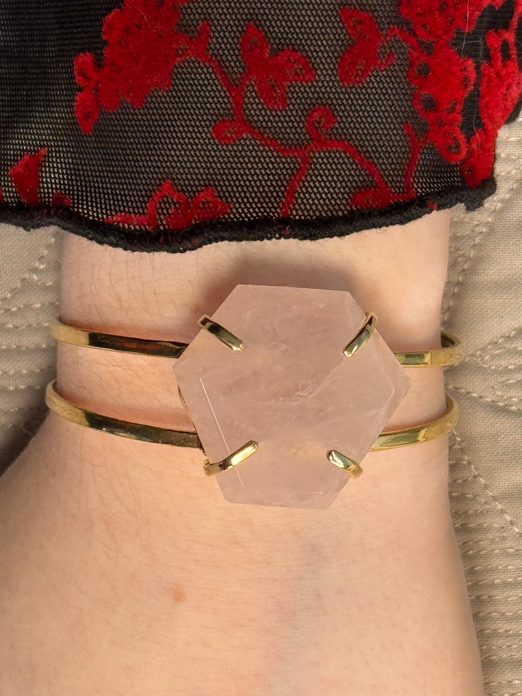Close-up of a wrist wearing a gold bracelet with a hexagonal pink stone on a textured surface.