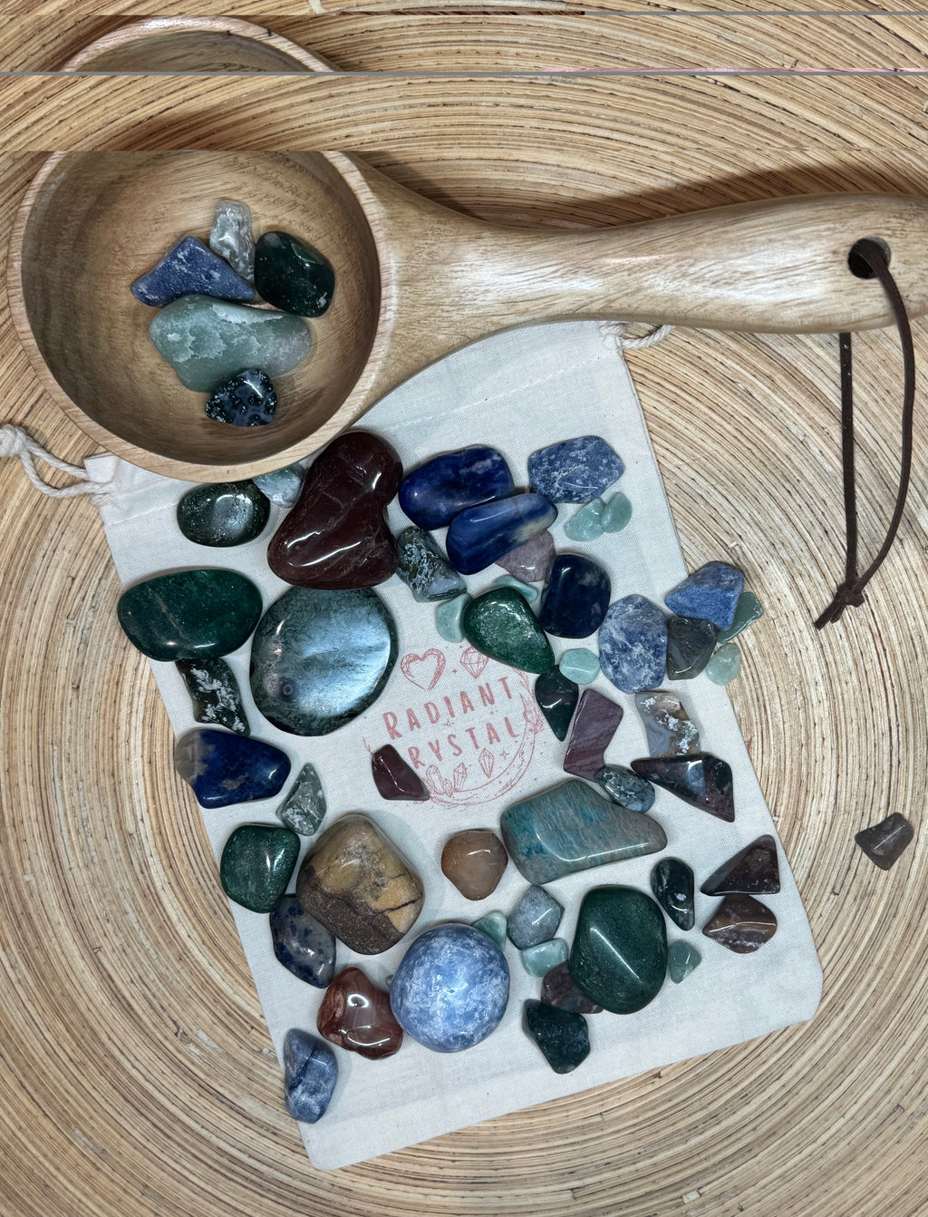 Assorted tumbled stones on a white drawstring bag with 'Radiant Crystal' branding, next to a wooden spoon on a wooden surface.